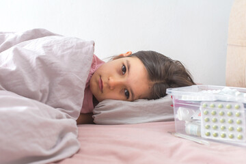 A sick child lying in bed under a pink blanket, resting her head on a pillow and looking tired. A...