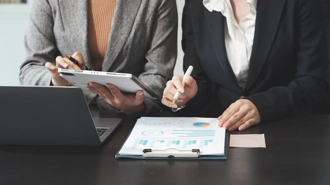 Two female business professionals in suits sit at desk, engaged in strategic discussion. They analyze financial planning, risk management, corporate growth while reviewing reports and market trends.
