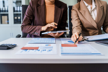 Two female business professionals in suits sit at desk, engaged in strategic discussion. They...