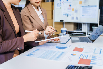 Two female business professionals in suits sit at desk, engaged in strategic discussion. They analyze financial planning, risk management, corporate growth while reviewing reports and market trends.