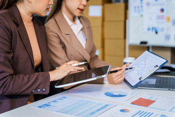 Two female business professionals in suits sit at desk, engaged in strategic discussion. They...