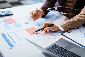 Two female business professionals in suits sit at desk, engaged in strategic discussion. They analyze financial planning, risk management, corporate growth while reviewing reports and market trends.