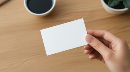 Obraz premium Blank Business Card in Hand on Wooden Table Next to Coffee and Small Plant for Personal Branding or Design