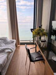Hotel room with a bed, a wooden desk, and a chair facing a panoramic sea view through a large window