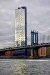 Manhattan Bridge with a glass skyscraper behind it reflected in the Hudson River