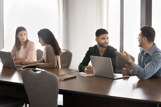 Team members engaged in pairs, discussing ideas in collaborative workspace, sit at shared table with laptops, brainstorm solutions, review project details, exchanging feedback to achieve common goal