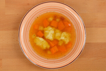 A top-down view of a bowl of clear soup with semolina dumplings and diced carrots, presented on a wooden surface