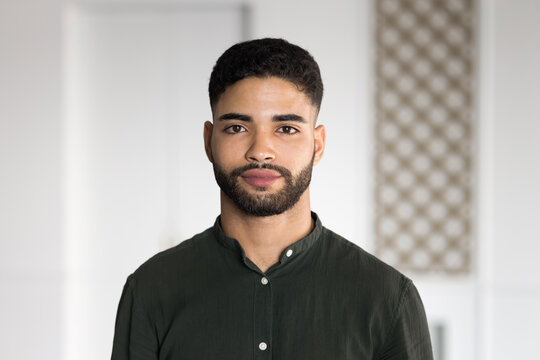 Young Arab man dressed in casual shirt standing indoors with serious expression, stare at camera, looks confident, head shot, office portrait. Human resources, employment, profile picture of applicant