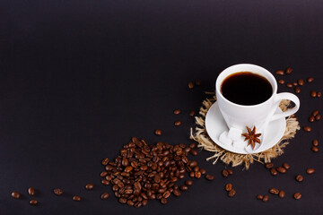 A full white cup of coffee sits on a saucer with sugar cubes and star anise. Coffee beans are scattered around the cup. The table is black