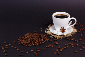 A white cup of black coffee sits on a saucer and jute mat with sugar cubes and star anise. Roasted coffee beans are scattered on a dark surface around it