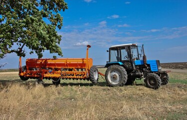 A blue tractor with a seeder stands on the edge of a field on a sunny day.