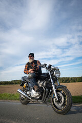 Fototapeta premium Cooler Motorradfahrer mit grauem Bart und Sonnenbrille präsentiert stolz sein klassisches Motorrad, Aufnahme im Hochformat.