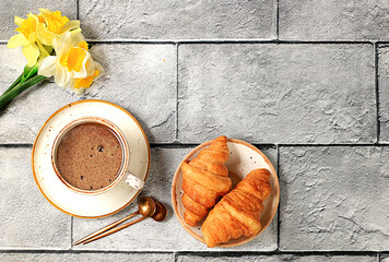 Delicious croissants, a cup of cappuccino and a bouquet of daffodils on a concrete table. Healthy breakfast with ingredients, the concept of a perfect morning and traditional national cuisine