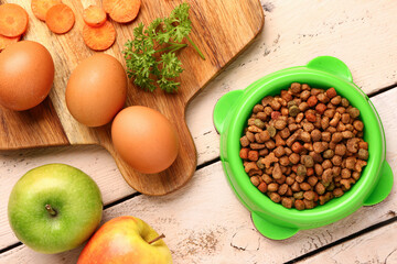 Bowl with dry pet food and natural products on light wooden background