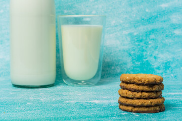 Stack of chocolate-covered oat cookies sits next to a full glass of milk, with a glass bottle visible in the background. Scene evokes warmth, comfort, and indulgence, perfect for food themes.