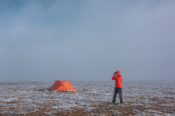 Man in red jacket in hood with photo camera near orange tent on snow-covered stony pass in sunlight...