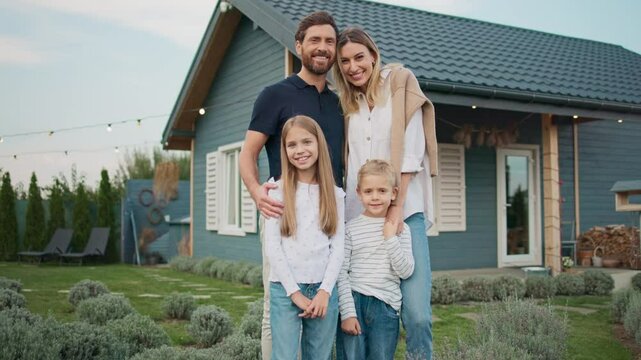 Caucasian husband and wife hugging their beloved kids. Family standing together in front of new building. Proud about newly purchased property. Small house with blue walls and green yard.