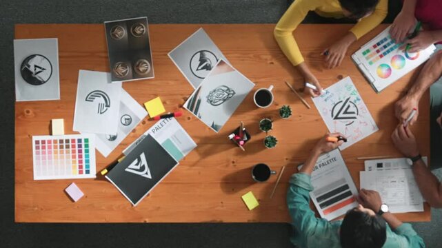 Top view of designer writing and designing logo graphic at meeting. Closeup of business man working on colored palettes and writing idea on sticky notes on table with equipment and paper. Symposium.