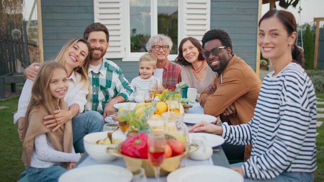 Cute family sitting together at table in front of cozy house. Having dinner in fresh air. Looking at camera while smiling. People having fun together. Tasty dishes on table. Evening in village.