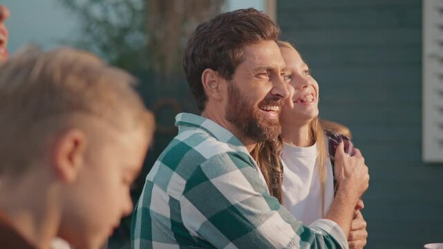 Side view of Caucasian father hugging his beloved child. Wearing checkered shirt. Smiling while showing love and care. Sun shining bright on peoples faces. Spending free time outside.