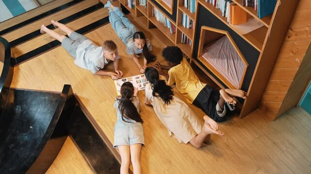 Group of diverse children lying down in circle while reading a book at library. Top view of girl sharing a magazine while pointing at interested topic and talking with lovely friends. Edification