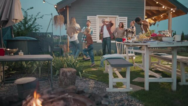 Camera moving closer to positive family. People spending time together. Celebrating something. Everyone dancing. Fire burning at fireplace. Table covered with many dishes. Evening.