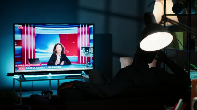 Relaxed person watching breaking news on television at night, chilling on sofa in living room and watching tv program. Young adult listening to media information on global network. Camera B.