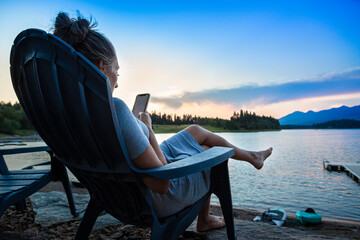 Woman relaxing from her lakeside home overlooking a scenic lake in the Montana mountains. She is sitting and looking at her smartphone with a beautiful sunset in the background.