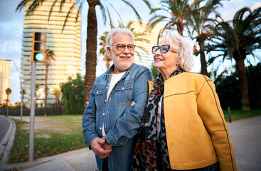An elderly couple stands together at a crosswalk, waiting for the traffic light to change. Their...