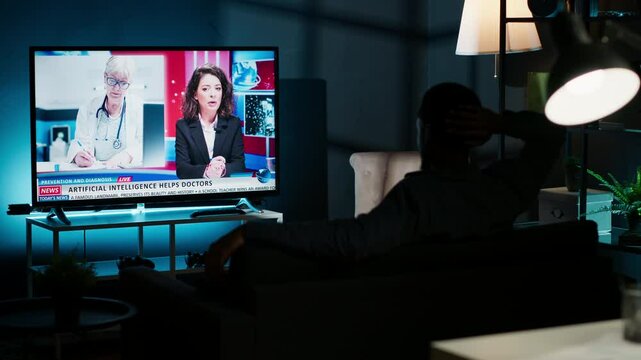 At night, young guy follows latest headlines on tv relaxing on couch in the living room, watching news program. On international network person pays attention to information broadcasted. Camera B.