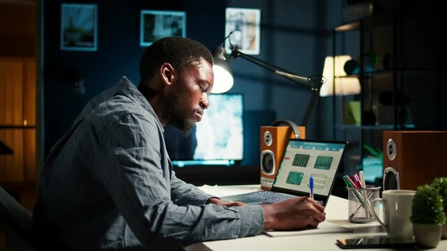 Self employed Black man multitasking at home taking handwritten notes and browsing the web for project research. Managing schedule and tasks with a planner to boost productivity. Camera A.