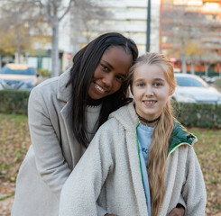 Young mother is hugging her Russian adopted daughter in an urban park, both are smiling and looking...