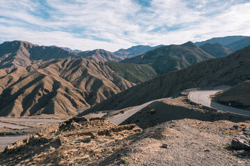 A winding road cuts through the rugged landscape of the Atlas Mountains in Morocco, under a partly cloudy sky.