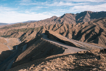 A winding road cuts through the rugged landscape of the Atlas Mountains in Morocco, under a partly cloudy sky.