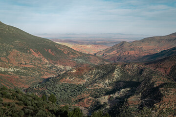 A panoramic view of the majestic Atlas Mountains in Morocco, contrasting against a clear blue sky. Lush green valleys wind through the rugged terrain,.