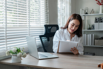 Businesswoman in a home office setting, writing on a clipboard with a laptop on the desk, surrounded by natural light.