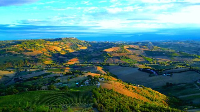 Vivid aerial pullback pan right shot from Montefiore dell'Aso (AP), striped fields, thickets, roads and buildings stand on sumptuous Marche hills, with balmy sunlight, dramatic cloudy sky, Sibillini