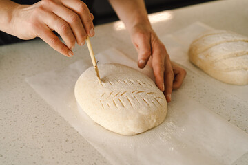 Woman hands skillfully score the surface of a round dough loaf, preparing sourdough bread for baking. The bright kitchen setting provides a warm atmosphere and fresh ingredients.