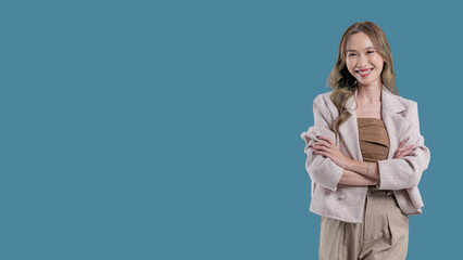 A woman is standing in front of a blue background, wearing a tan jacket and tan pants. She is smiling and she is confident