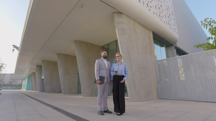 A confident businessman and a dynamic businesswoman stand outside a modern glass building, discussing their latest ventures, collaborative strategies, and innovative ideas for success