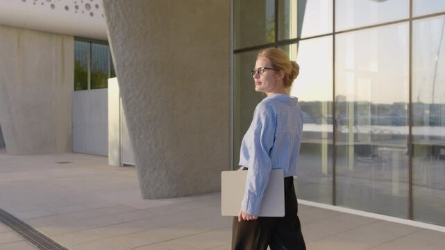 A confident professional woman wearing glasses walks down a busy city street, balancing a sleek laptop. Surrounded by stunning modern architecture that merges aesthetics with practical functionality