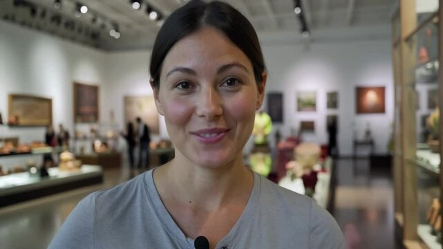 A Smiling Young Woman in a Light Gray T-Shirt Inside an Art Gallery with Colorful Artwork and Visitors in the Background