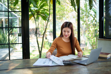 Businesswoman working on a laptop with documents in a natural-themed office, focused and determined...