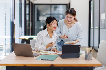 Fototapeta premium Two smiling happy asian businesswomen working together on a laptop in the office, demonstrating teamwork business meeting in a corporate and collaboration 