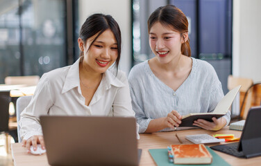 Happy professional young women collaborating with enthusiasm in a modern office setting. Two asian female entrepreneurs using a laptop for a productive business meeting in a corporate workplace.
