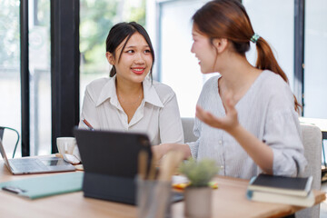 Happy professional young women collaborating with enthusiasm in a modern office setting. Two asian female entrepreneurs using a laptop for a productive business meeting in a corporate workplace.
