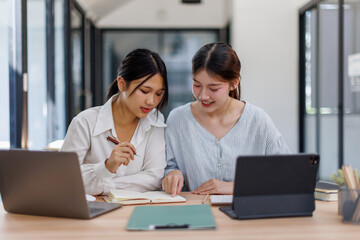 Happy professional young women collaborating with enthusiasm in a modern office setting. Two asian female entrepreneurs using a laptop for a productive business meeting in a corporate workplace.
