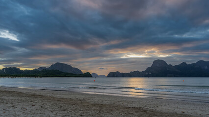 Evening after sunset on a tropical island. Pastel colors. The blue clouds in the sky are highlighted in pink. Silhouettes of mountains on the horizon. Glare, reflection on the surface of a calm ocean
