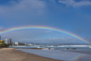 A rainbow, representing the calm before the storm, curves over a beach with rough surf driven by an approaching cyclone or hurricane edge at Coolangatta on the Gold Coast in Queensland, Australia.