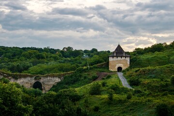 Historic medieval fortress with a stone entrance gate surrounded by lush greenery. Scenic landscape with an old arched bridge, perfect for travel and history-related themes. "Khotyn Fortress"